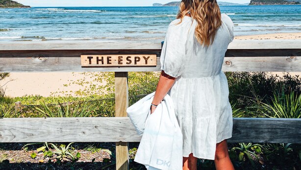 Eine Frau in weißem Kleid und Strohhut steht auf einem Holzboden und blickt auf einen Strand. Ein Schild zeigt "THE ESPY".