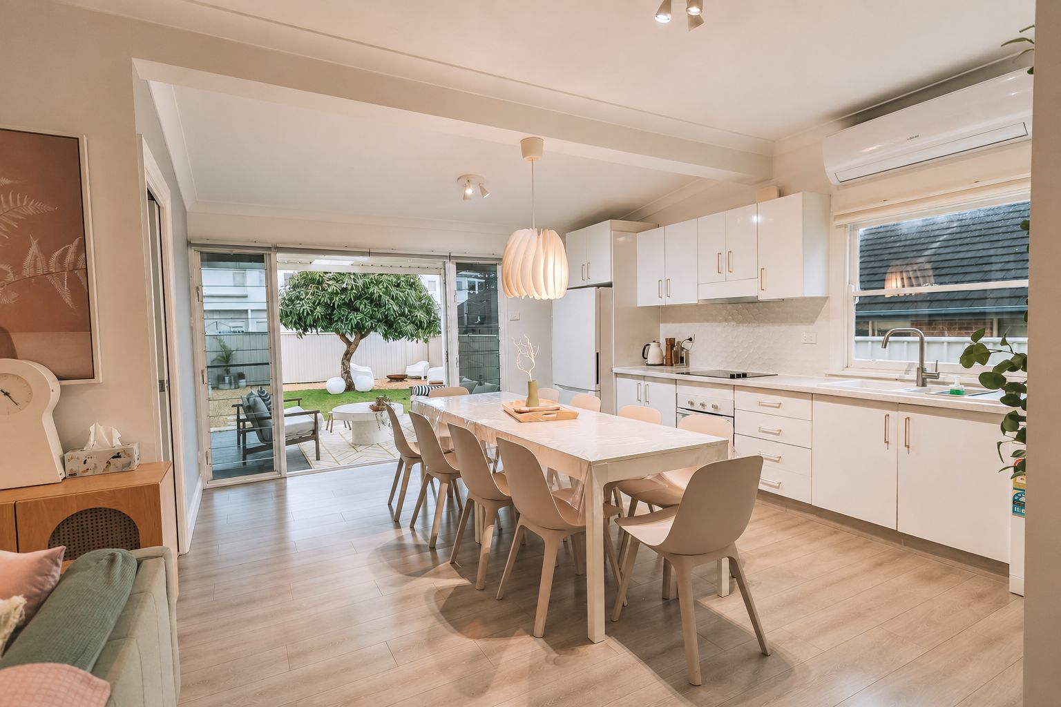 A modern kitchen with white cabinetry, a dining area with a table and chairs, and a view of a tree through a glass door.