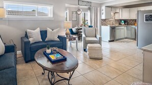 A modern living room with a blue sofa, a coffee table, and a dining area with a chair and table. The kitchen is visible with cabinets and a refrigerator. - Great Location!! Family Friendly, 3 Pools, Lake, Tennis, Free Activities & Walk to Restaurants/Shops (Destin)