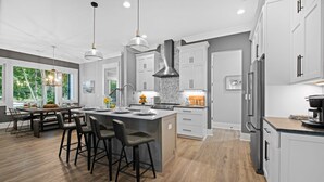 A modern kitchen with a central island, bar stools, and a dining area with a view of the outdoors. - Hanesbee Escape w/Beach, Hot Tub & Optional Boat Rental (Mooresville)