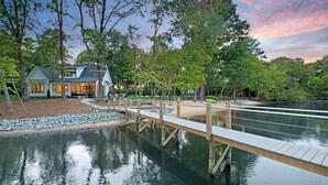 A lakeside property with a wooden dock, a house with a landscaped yard, and a calm lake. - Hanesbee Escape w/Beach, Hot Tub & Optional Boat Rental (Mooresville)