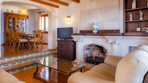Uma sala de estar com lareira, vigas de madeira, TV, área de refeições com cadeiras e escada com azulejos estampados.