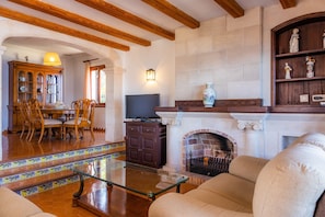 A living room with a fireplace, wooden beams, a TV, a dining area with chairs, and a staircase with patterned tiles.
