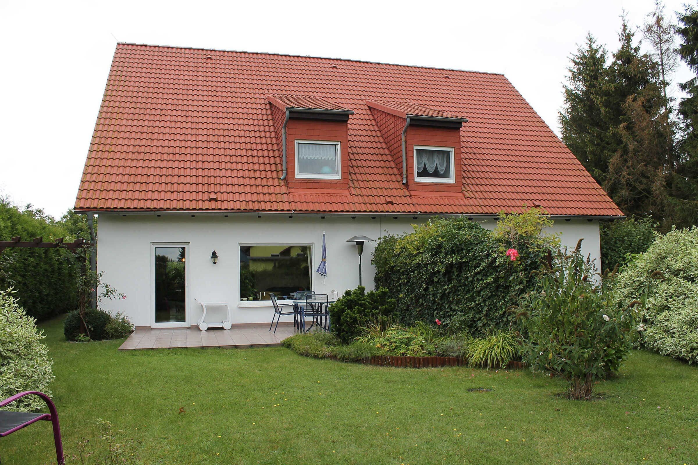 A house with a red roof, a front door, a patio with a table and chairs, and a garden with various plants.