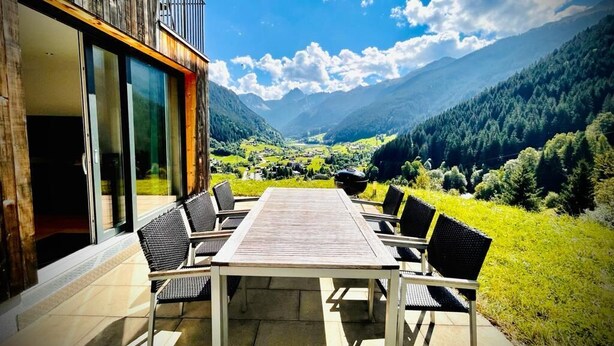 A wooden building with a balcony, a table and chairs outside, and a view of a valley and mountains. - Modern, idyllic vacation home with breathtaking panoramic views (Gaschurn)