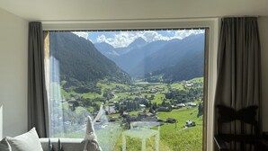 A room with a large window offering a view of a valley with houses and mountains. - Modern, idyllic vacation home with breathtaking panoramic views (Gaschurn)