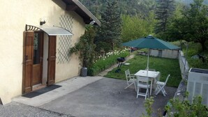 Un patio con tavolo e sedie sotto un ombrellone verde, una porta in legno e un giardino con fiori e alberi.