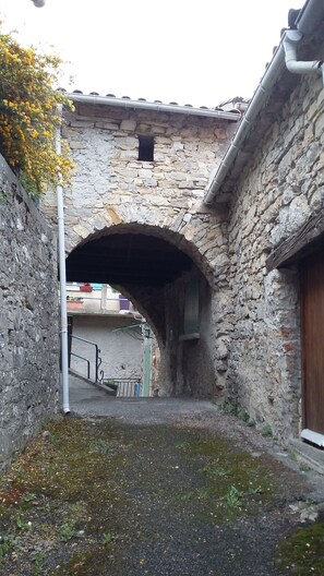 Ein steinerer Bogen führt zu einem Innenhof, an dessen Seite eine Steinmauer und auf der anderen Seite ein Gebäudeeingang zu sehen sind.