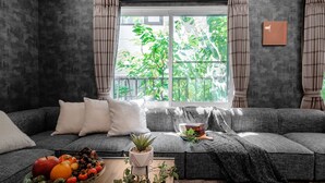 Una sala de estar moderna con un sofá gris, una mesa de centro de madera con frutas y una planta en maceta, y un ventanal con cortinas.