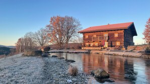 A wooden cabin with a red roof, a balcony, and a stone wall. - Vacation home rehberg14 - historic farm with pond & sauna (Drachselsried)
