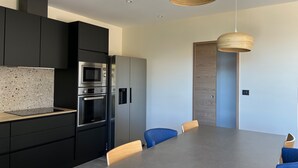 Une cuisine moderne avec des meubles noirs, un four encastré et un réfrigérateur en acier inoxydable. On y trouve une table à manger avec des chaises bleues et un luminaire en bois suspendu au plafond.