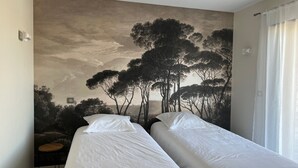Une chambre avec deux lits, une fresque représentant des arbres, une petite table et une fenêtre avec des rideaux.
