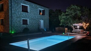 Una piscina con bordo protetto da una rete, circondata da un edificio in pietra con numerose finestre e un'area salotto esterna coperta con luci decorative.
