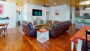 A spacious living area with a wooden ceiling, a flat-screen TV, a large sofa, and a kitchen area with green chairs. - Near Beach, Balcony Overlooking Private Pond, Private Pool (Port Aransas)