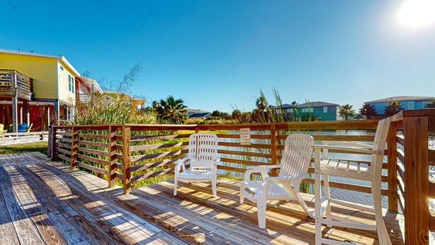 A wooden deck with white chairs, a wooden fence, and a sign warning against casting toward the fountain. - Near Beach, Balcony Overlooking Private Pond, Private Pool (Port Aransas)