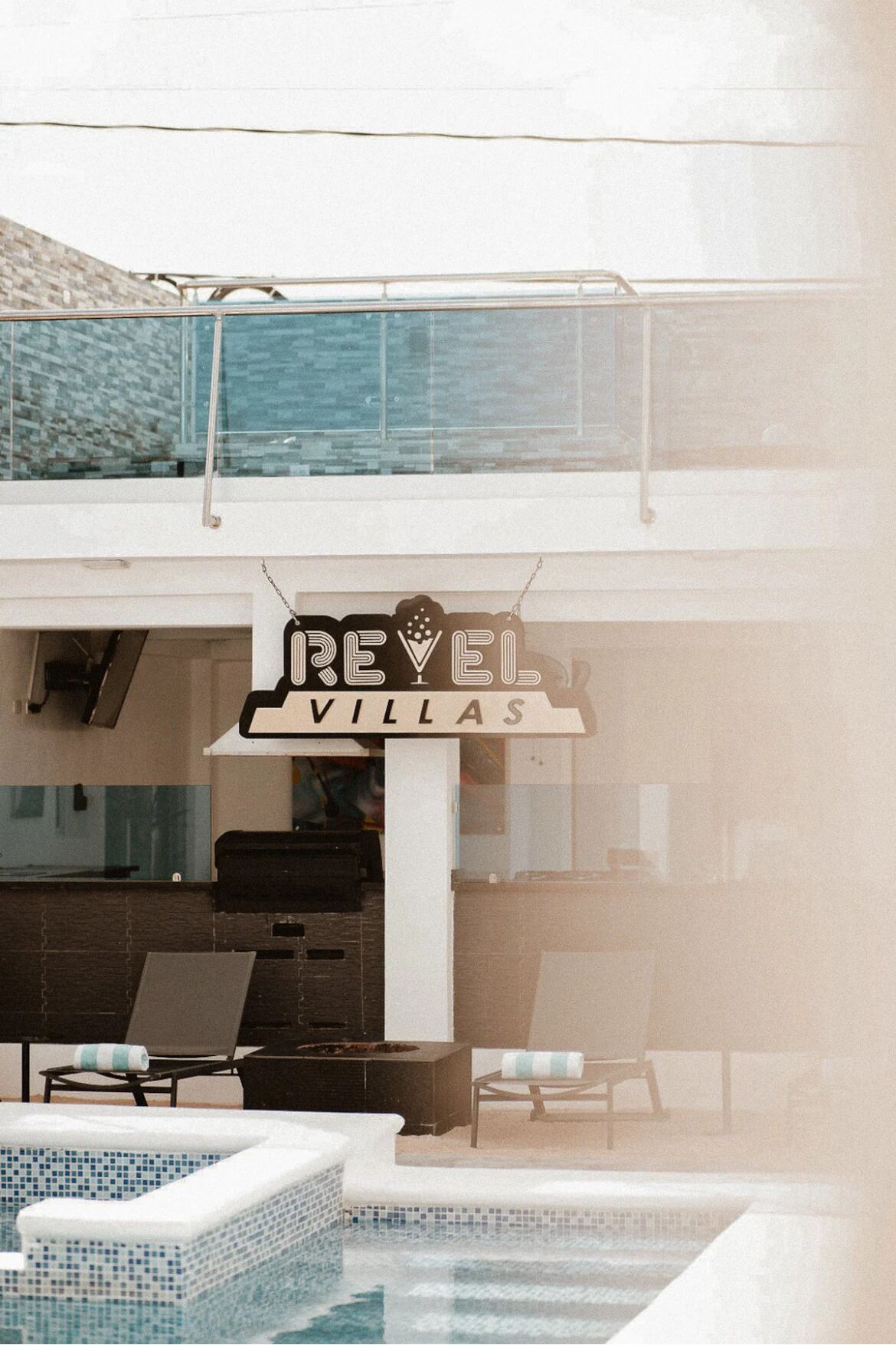 Une enseigne indiquant « REVEL VILLAS » au-dessus d’un espace piscine équipé de chaises longues et offrant une vue dégagée sur la piscine.