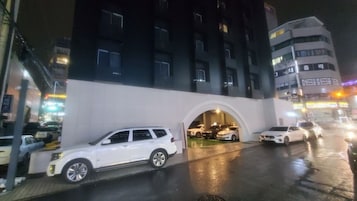 Un edificio de varios pisos con fachada blanca y oscura, una zona de estacionamiento techada y varios autos estacionados al frente.