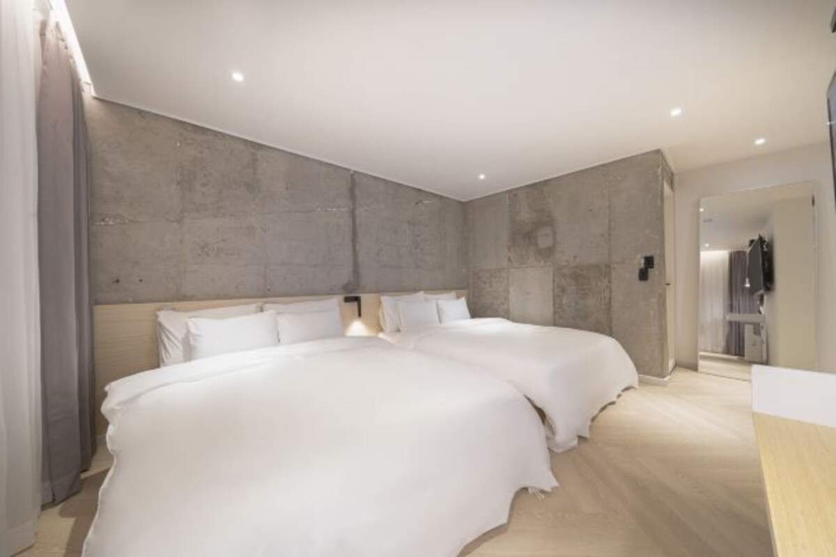 A modern hotel room with two large beds, a concrete accent wall, and a wooden floor.