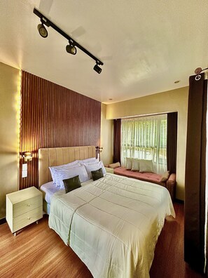 A hotel room with a large bed, a bedside table, a nightstand, a window with curtains, and a wall-mounted light fixture.