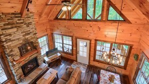 Une cabane avec une cheminée en pierre, des murs en bois, un ventilateur de plafond et de grandes fenêtres.