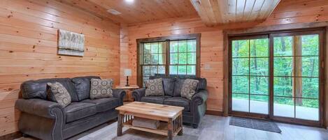 A log cabin interior with two sofas, a coffee table, and large windows offering a forest view.
