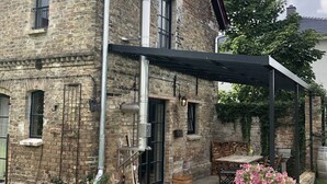 A stone house with a covered patio, a brick chimney, and a small garden with pink flowers. - Vacation apartment Beelitz - Barn Zauchwitz (Beelitz)
