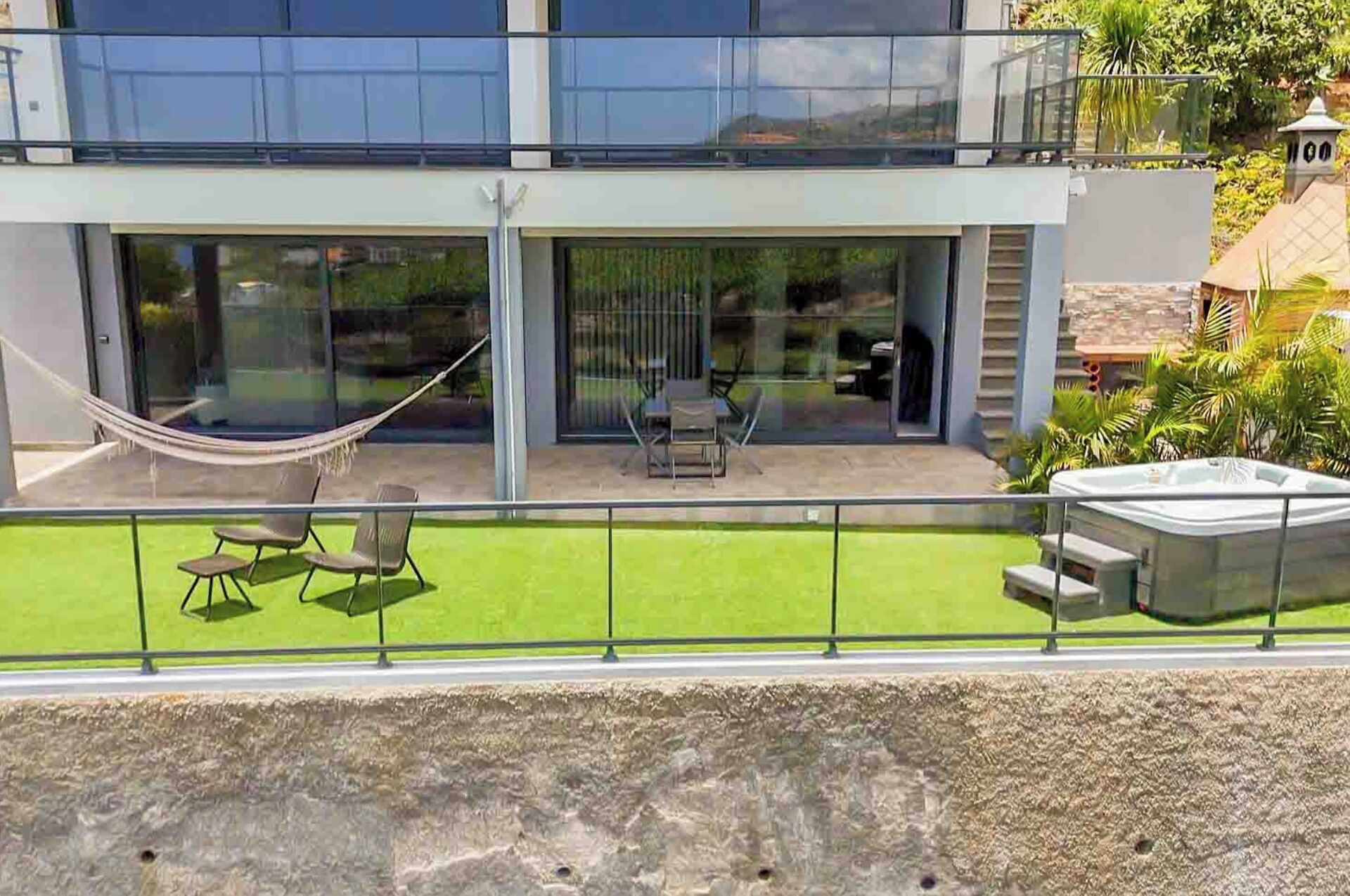 A modern two-story house with a glass facade, a hammock, outdoor furniture, and a hot tub.