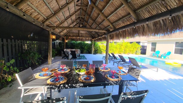An outdoor dining area with a thatched roof, surrounded by a pool and greenery. - POSHPADZ Presents Villa Tranquila- Private Pool & Tiki Close to Beach Sleeps 10 (Jupiter)