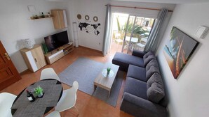 Un salon moderne avec un canapé gris, un téléviseur, une table à manger avec des chaises et une petite table basse. Un planisphère est accroché au mur, ainsi qu’un tableau représentant une scène de plage.
