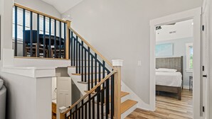 A staircase with wooden steps and black railings leading to a bedroom with a bed and a nightstand. - Landon Sea: Private Pool, Hot Tub, Dog-Friendly (Corolla)