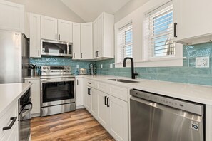 A modern kitchen with white cabinets, stainless steel appliances, a dishwasher, and a window with blinds. - Landon Sea: Private Pool, Hot Tub, Dog-Friendly (Corolla)