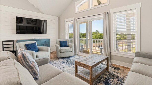 A spacious living room with a large window, a ceiling fan, and a television mounted on the wall. - Landon Sea: Private Pool, Hot Tub, Dog-Friendly (Corolla)