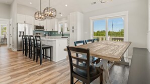A modern kitchen with white cabinets, a wooden dining table, and a view of greenery outside. - Landon Sea: Private Pool, Hot Tub, Dog-Friendly (Corolla)