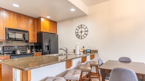 A modern kitchen with wooden cabinets, a stainless steel refrigerator, a microwave, and a built-in oven. There is a large wall clock and a bar stool area with a wooden table and chairs.