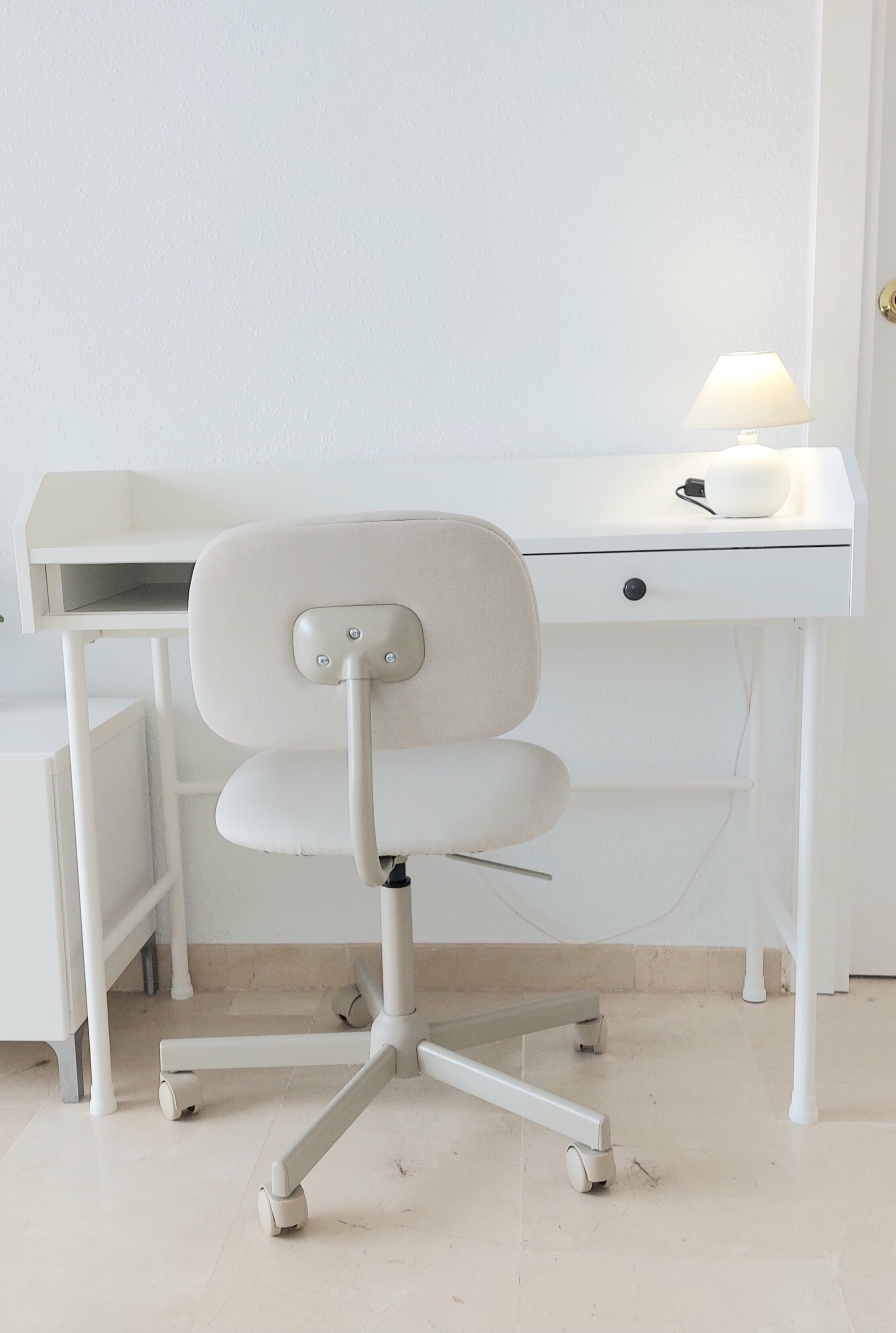 A white desk with a lamp, a chair with wheels, and a drawer.