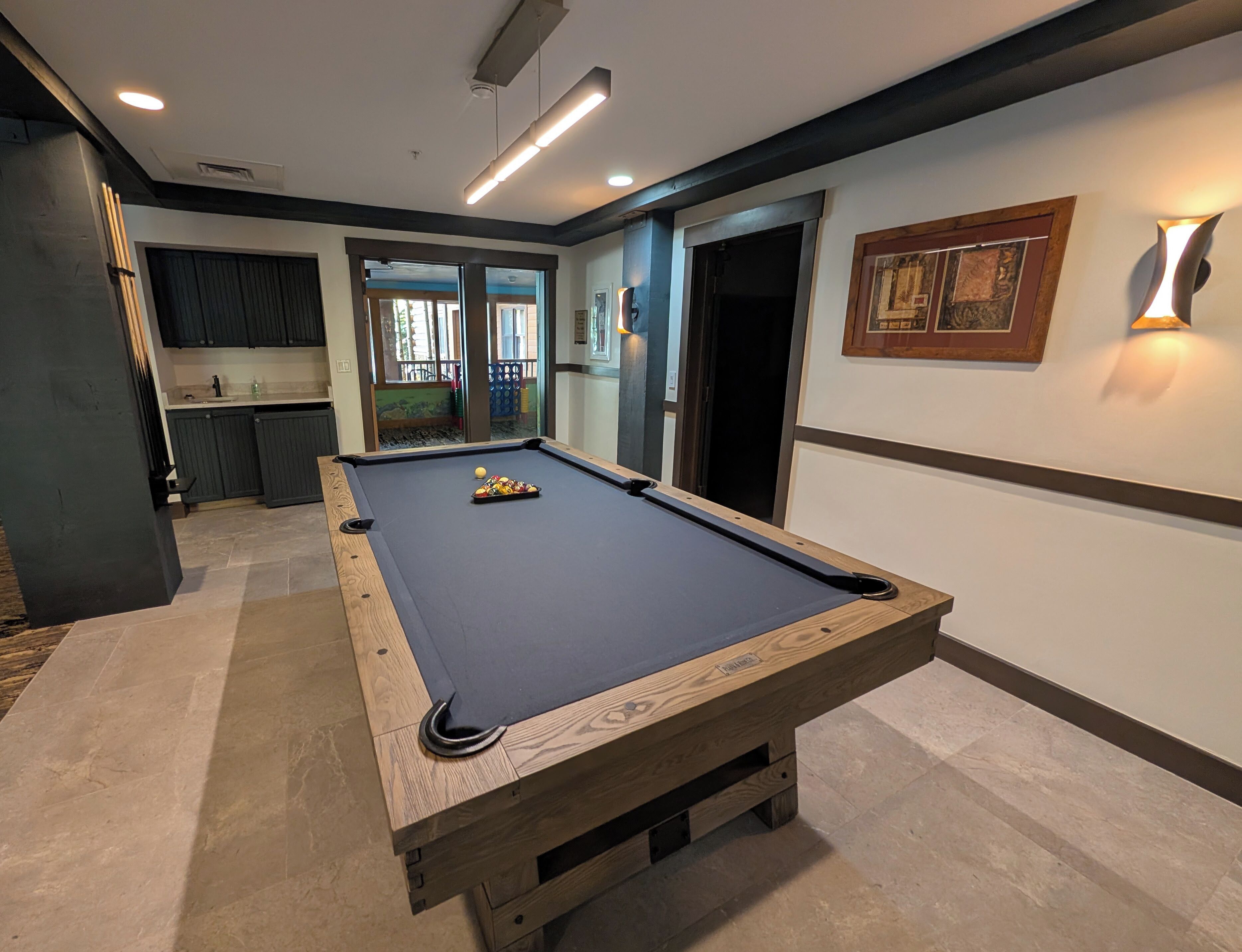 Une table de billard dans une pièce donnant sur l’extérieur, un coin cuisine et un tableau encadré au mur.