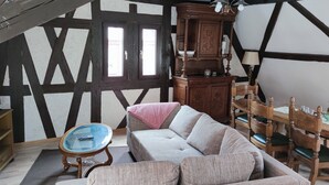 Een kamer met een hoge plafond, zichtbare balken, een houten eettafel met stoelen, een bank, een glazen bijzettafeltje en een kast.