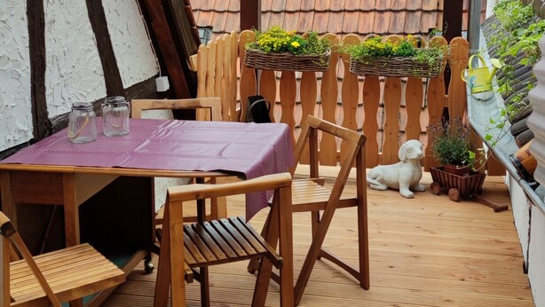 Een overdekt terras met houten meubels, een tafel met een paarse tafelkleed en potplanten.