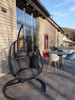 Un espace extérieur moderne, aménagé avec des sièges, comprenant un fauteuil suspendu, une table avec des chaises et une vue sur un bâtiment doté de grandes fenêtres.