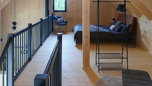 Ein Schlafzimmer im Loft-Stil mit Holzbalkendecke, einem Bett, einem Sessel, einem kleinen Tisch und einem Fenster mit Blick auf Bäume.