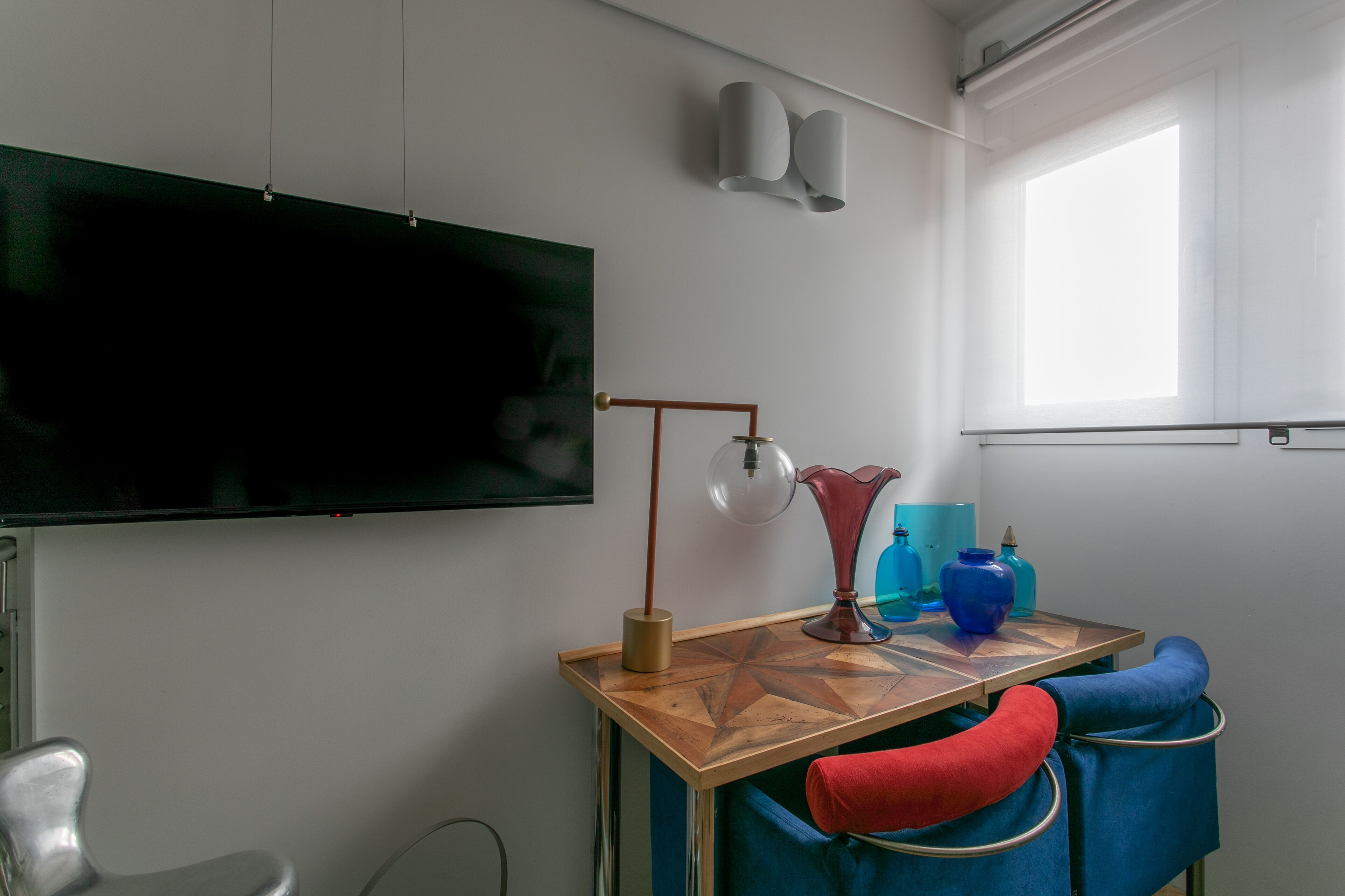 Una habitación con una televisión de pantalla grande montada en la pared, una mesa de madera con una lámpara y una ventana que proporciona luz natural.