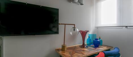 Una habitación con una televisión de pantalla grande montada en la pared, una mesa de madera con una lámpara y una ventana que proporciona luz natural.