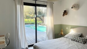 Une chambre à coucher comprenant un lit, une table de chevet, une lampe, une fenêtre avec des rideaux et une vue sur une piscine.