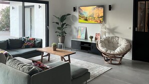 Un salon moderne avec un canapé d’angle, une table basse en bois et un fauteuil en rotin. Un grand téléviseur à écran plat est fixé au mur, ainsi qu’une plante en pot et un élément décoratif mural.