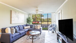 A living room with a sectional sofa, a coffee table, a flat-screen TV, and a large window with a view of palm trees and a road.