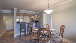 A dining area with a round table and six chairs, a kitchen in the background, and a chandelier.