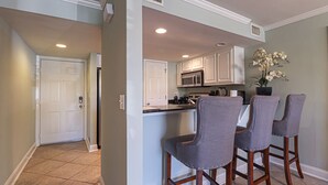A kitchen with white cabinets, a microwave, and a sink. There are three bar stools with a gray fabric seat and wooden legs.