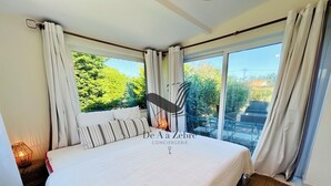 A hotel room with a large bed, a view of greenery outside, and a dining area visible through the window.