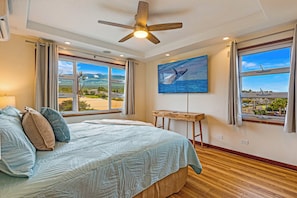 A bedroom with a bed, a ceiling fan, a painting of a whale, and large windows with a view.