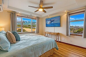 Un dormitorio con cama, ventilador de techo, un cuadro de una ballena y ventanas amplias con vistas.